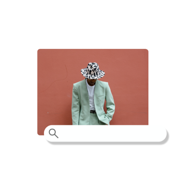 Eine Person im mintgrünen Anzug und schwarz-weißem Hut steht vor einer rostroten Wand.
